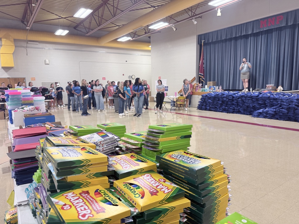 A large room with stacks of boxes, people gathered, a stage with a person speaking, blue curtains, and yellow ceiling lights.