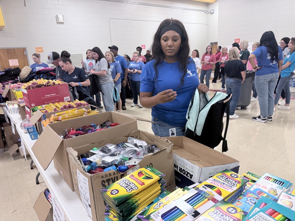 A woman in a blue shirt holds a backpack near boxes of crayons, pencils, and other school supplies.