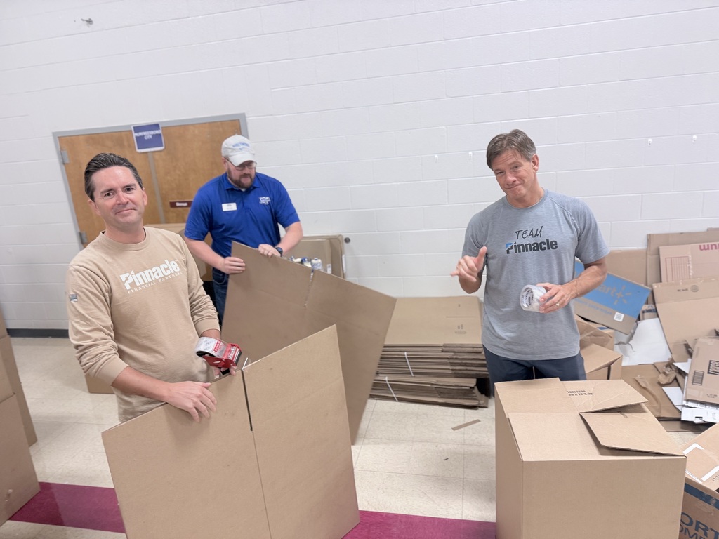 Three men in a room, holding cardboard boxes. One holds a box in front, another holds a box behind, and a third holds a roll of tape.