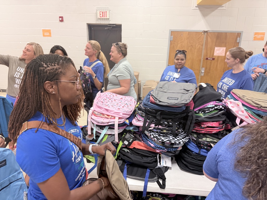 People in blue shirts gather around a table. Many bags, including backpacks, are stacked on the table.