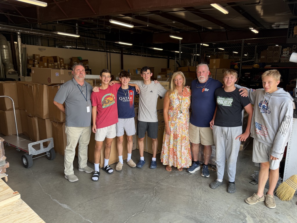 Group of people in a warehouse with boxes, standing together, smiling, and some holding onto shoulders.