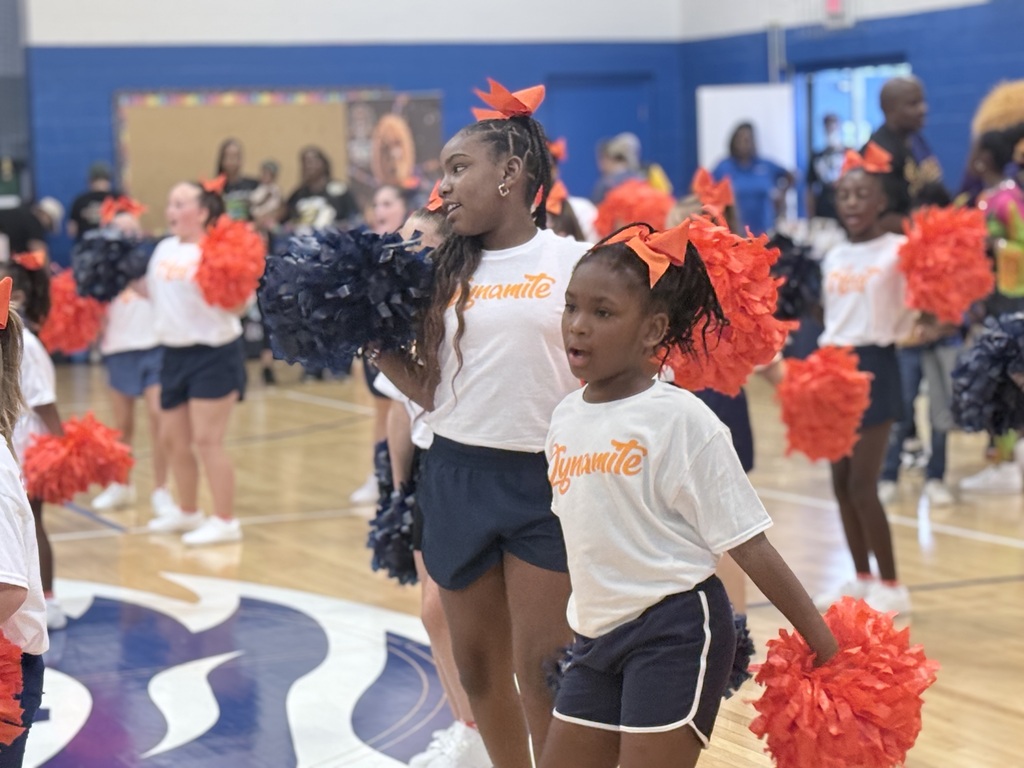 A group of young cheerleaders, two in front holding pom-poms, wearing white shirts and black shorts, in an indoor basketball court.
