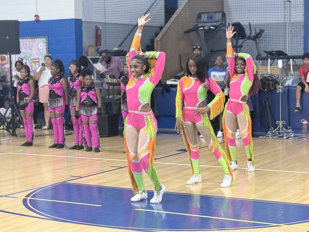 A group of women in colorful costumes dance on a basketball court, with others watching in the background.