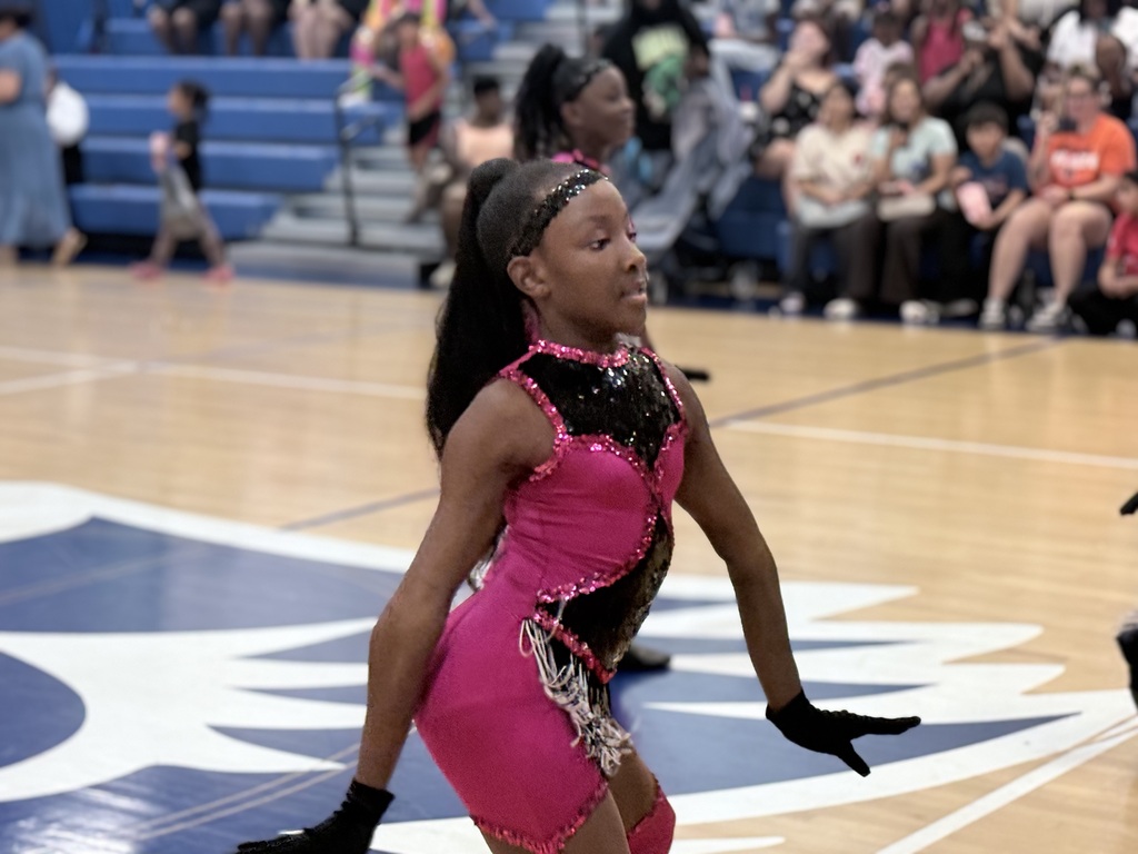 A young girl performs on a basketball court, wearing a pink and black costume. An audience sits in bleachers.