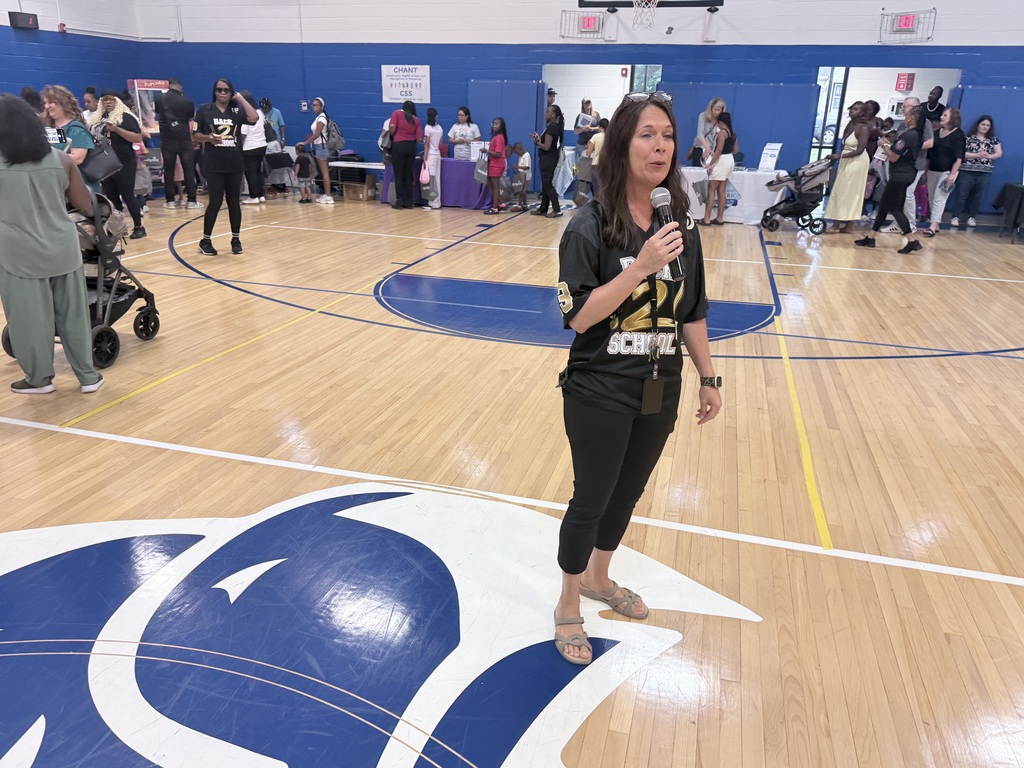 A person addresses a crowd in an indoor basketball court. A blue and white logo is on the floor.