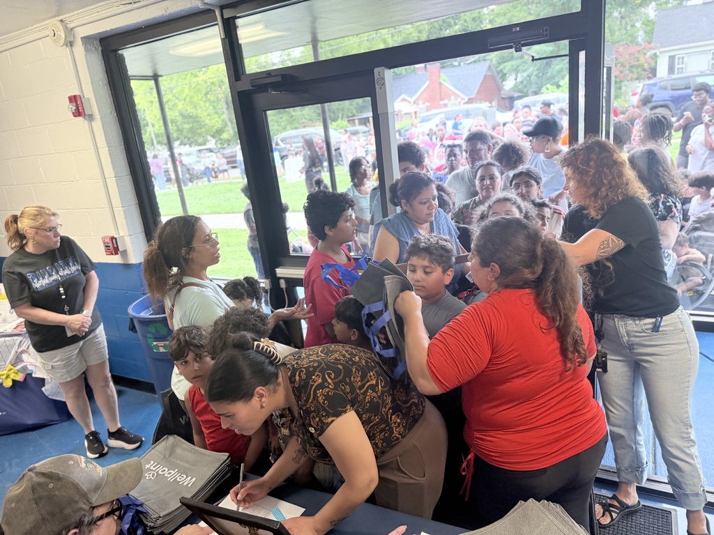 People are gathered indoors around a table. A woman in a red shirt and a hat assists a child.