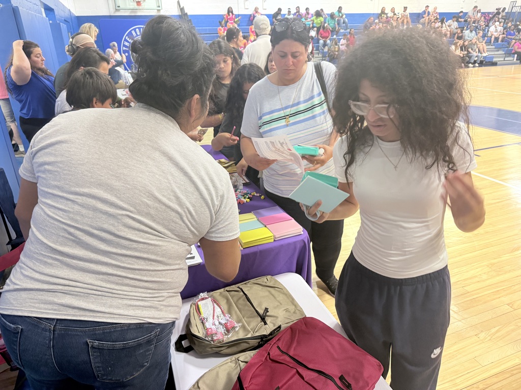 People gathered in a gymnasium. Two women with curly hair stand by a table with backpacks, books, and a purple tablecloth.