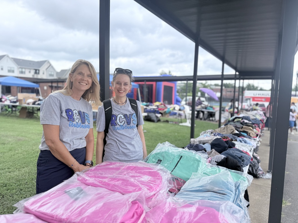 Two women stand under a canopy, smiling, near a table with many backpacks and bags. Behind them is a grassy area with tents.