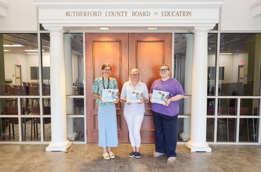 Three people stand in front of a wooden double-door, each holding a certificate. Behind them, large glass panels and white columns create an open, bright space.