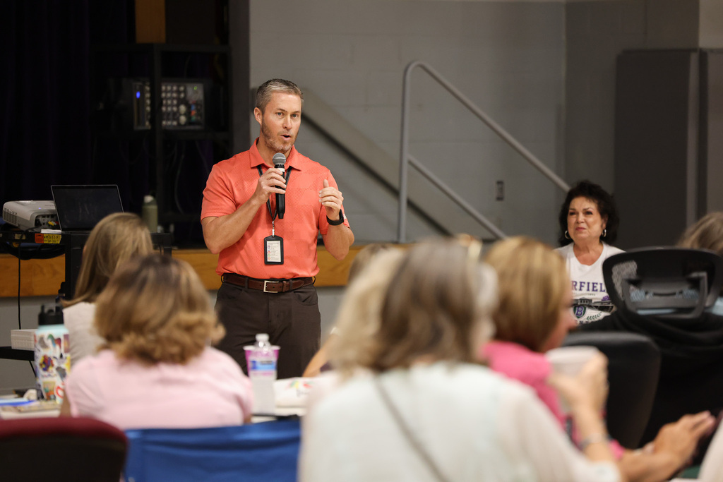 A man in an orange shirt stands at a podium, addressing a group of seated women in a classroom.