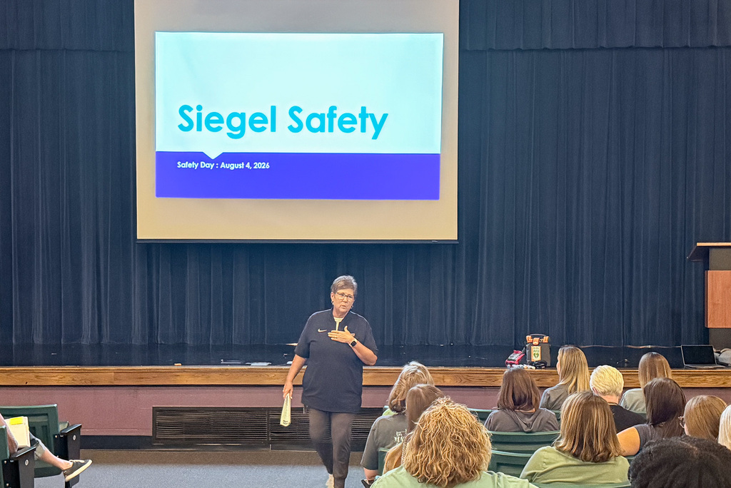 Person in black shirt walking past seated people, in front of a screen displaying "Siegel Safety" text.