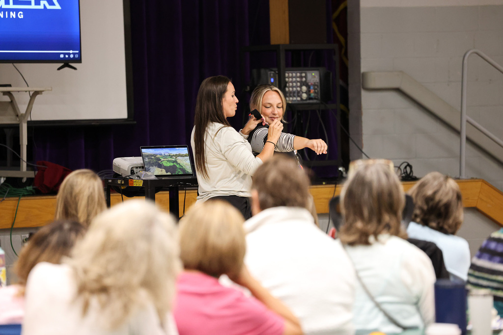 Two women stand before an audience, one speaking into a microphone. Behind them, a screen displays text.