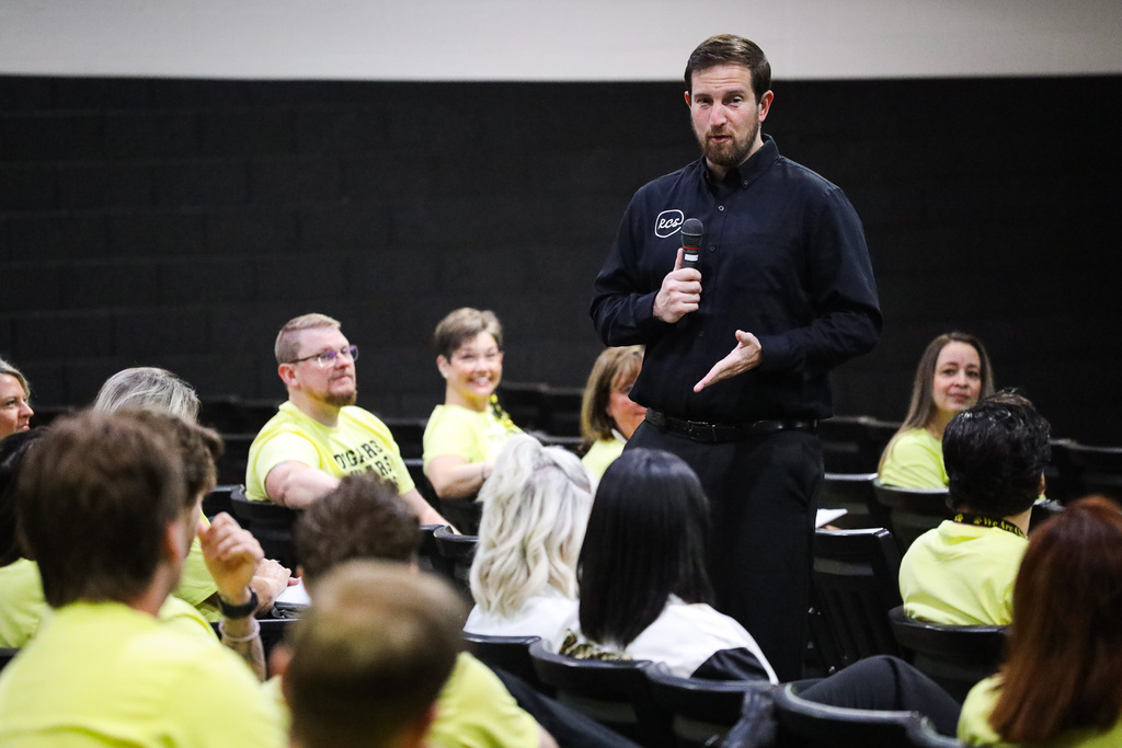 A man in a black shirt stands before a seated audience in yellow shirts, holding a microphone and gesturing.