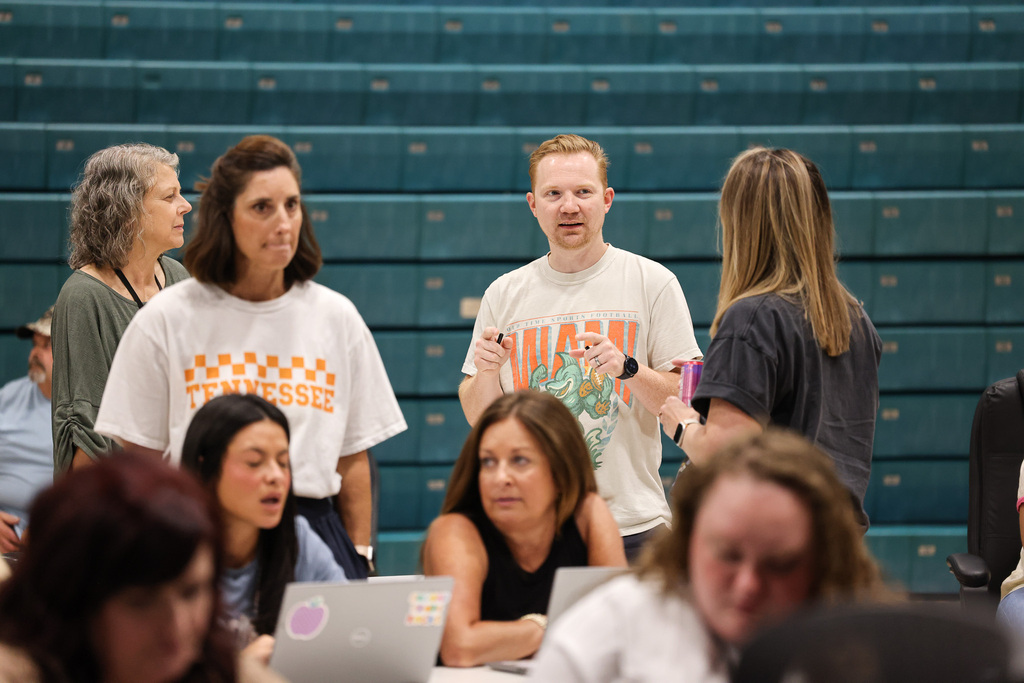 A group of people gathered around laptops, some standing, in front of a bleacher with a teal background.