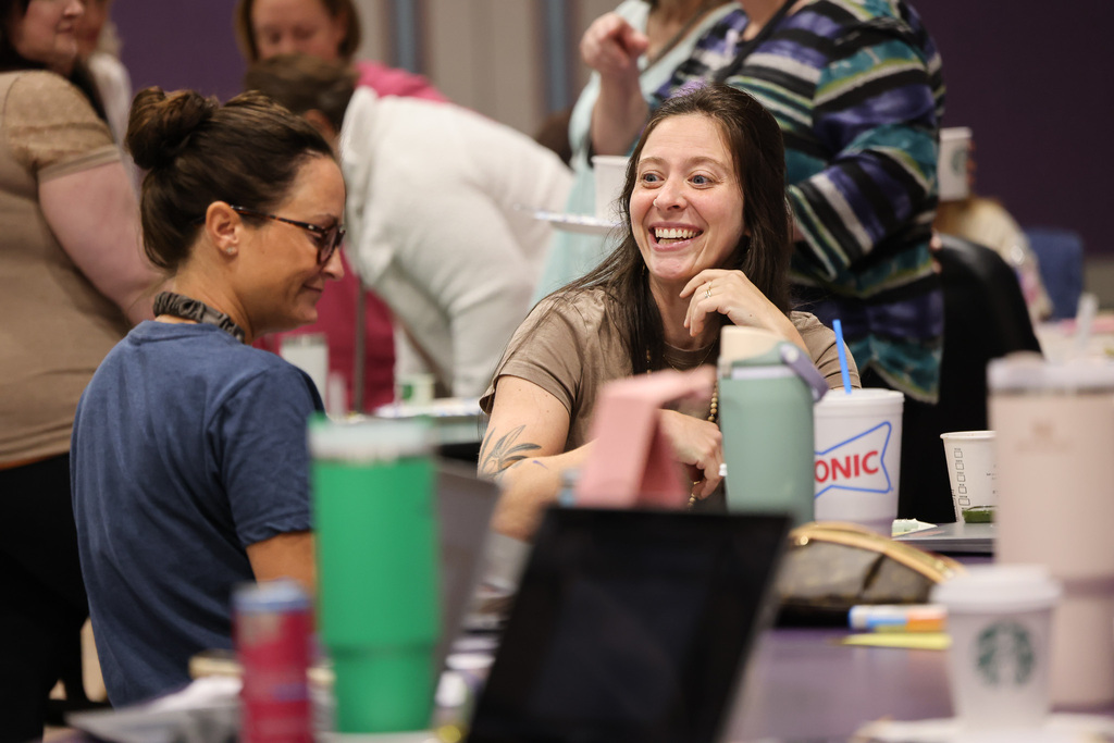 Two women converse with smiles; one wears glasses, and the other has a tattoo. Nearby, cups, a laptop, and a smartphone are on a table.