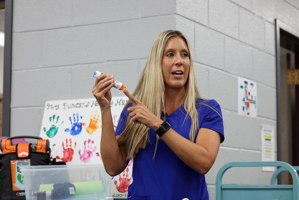 A woman in a blue shirt holds a marker and points at something while speaking, in a room with a white wall.