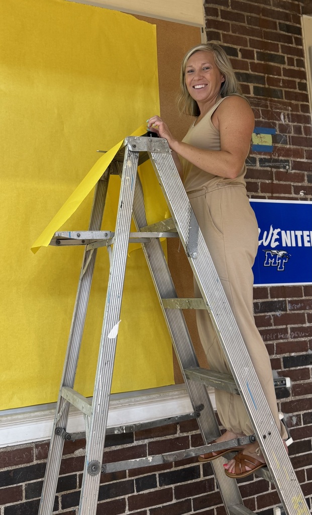 A woman stands on a ladder, holding yellow paper against a yellow wall, wearing sandals and a jumpsuit.