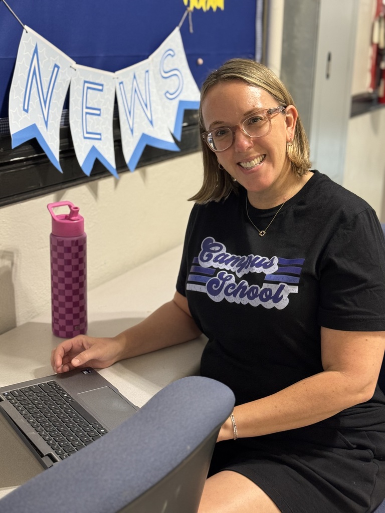 Woman in a black Campus School shirt seated at a desk with a laptop, water bottle, and a news banner behind.
