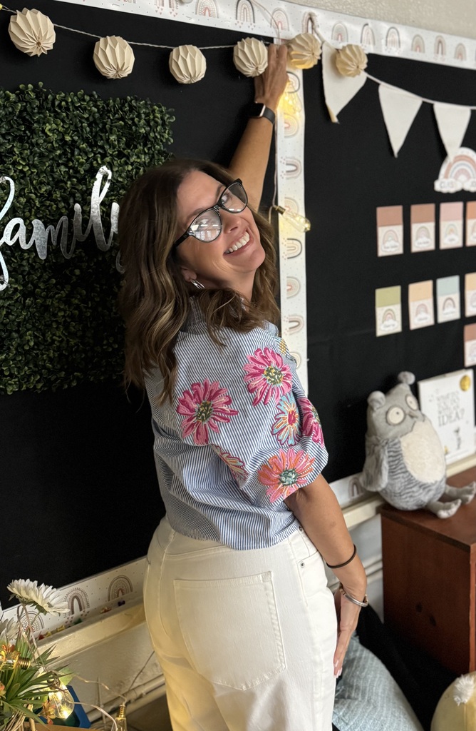 A woman wearing glasses and a floral top stands near a bulletin board, smiling. The board has colorful decorations.