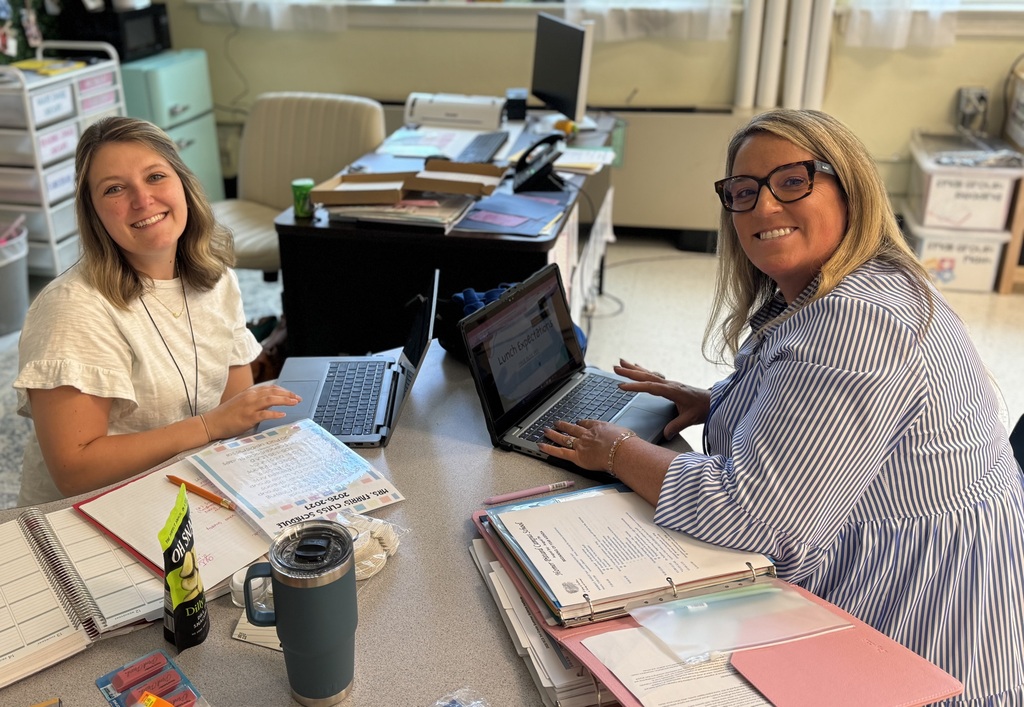 Two women sit at a desk with laptops, books, and office supplies. One wears glasses, the other a necklace.