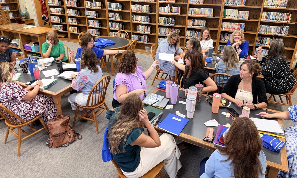 A group of women in a library, seated around tables with books, binders, and bags.