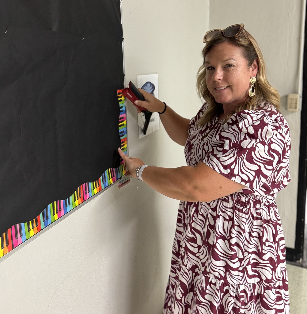 A woman in a floral dress stands next to a bulletin board with colorful piano keys.