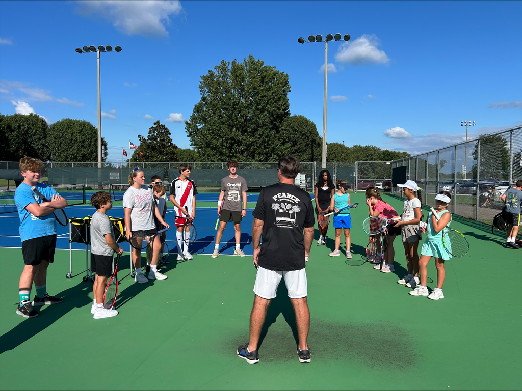 A group of children gather around a man on a tennis court, holding rackets and looking at him.