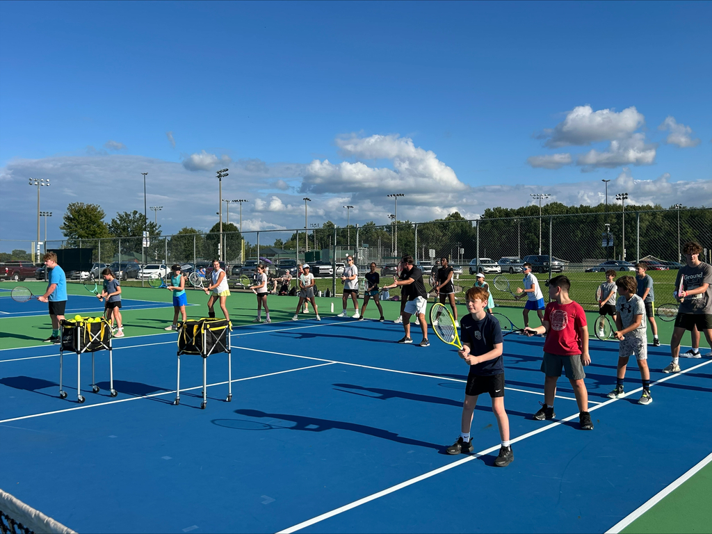 Group of people on a tennis court. Blue surface, white lines, trees and parked cars in the background.