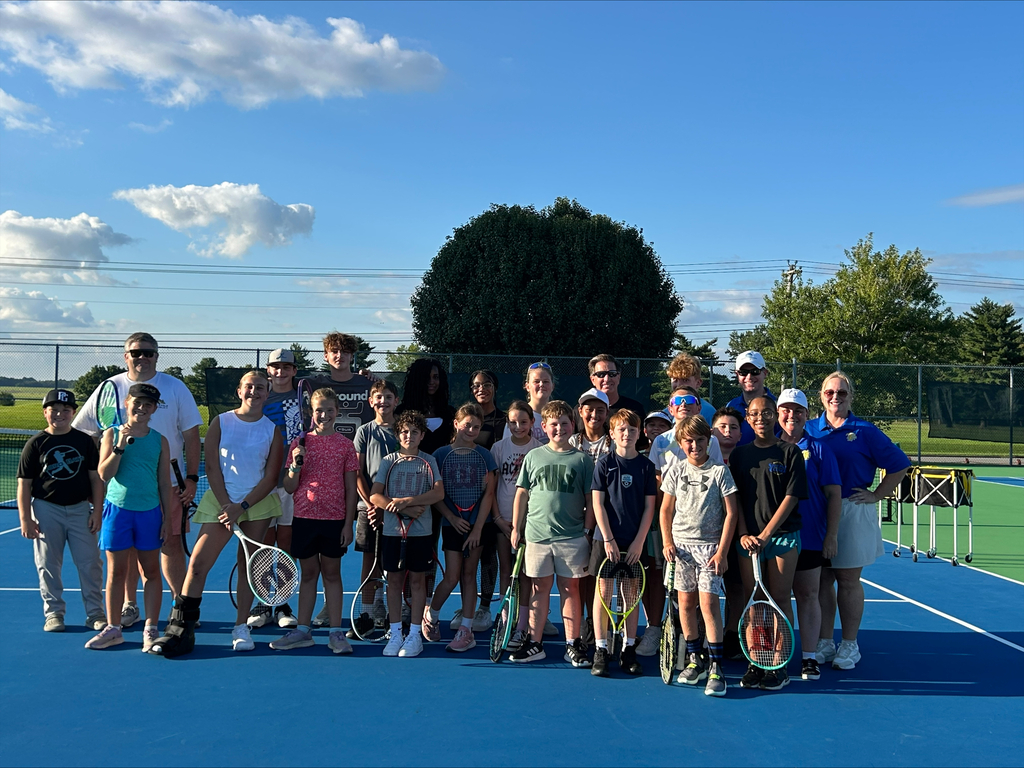 A group of people pose for a picture on a tennis court, holding rackets, with a tree and fence in the background.