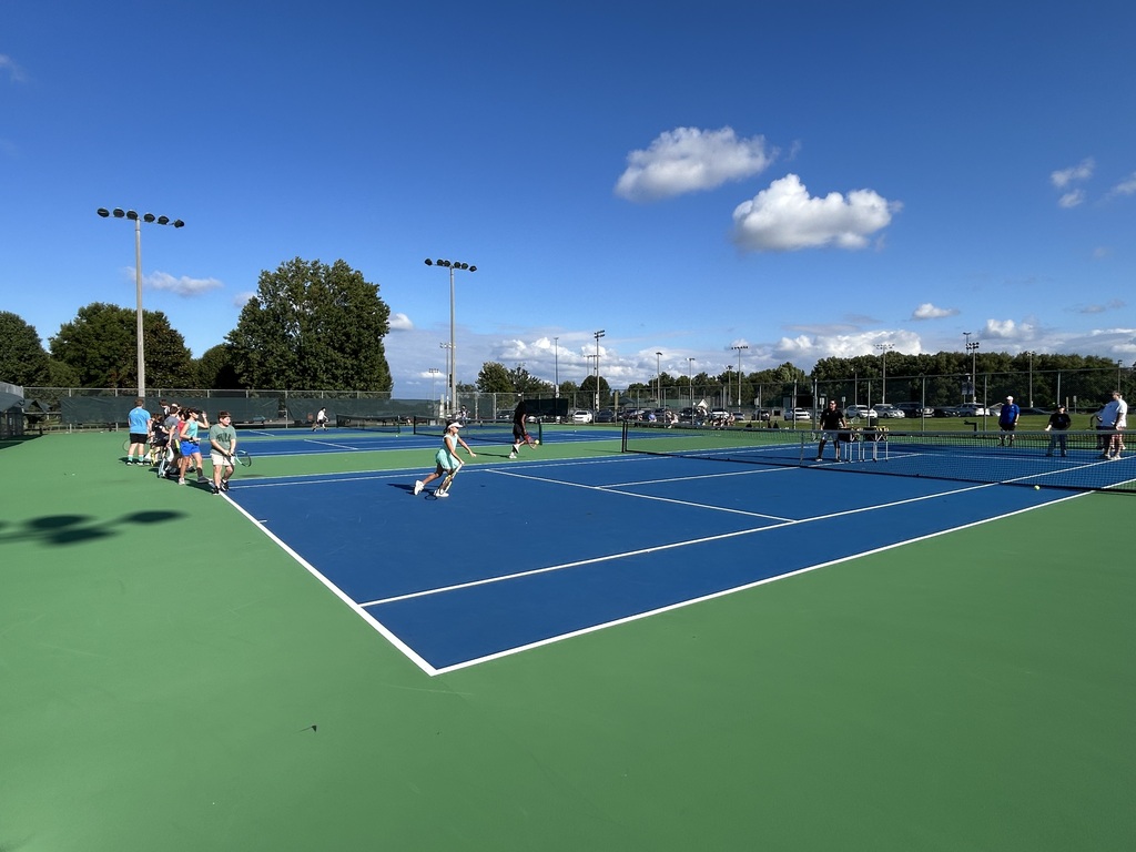 Multiple tennis courts with people playing tennis. A clear blue sky with a few clouds.