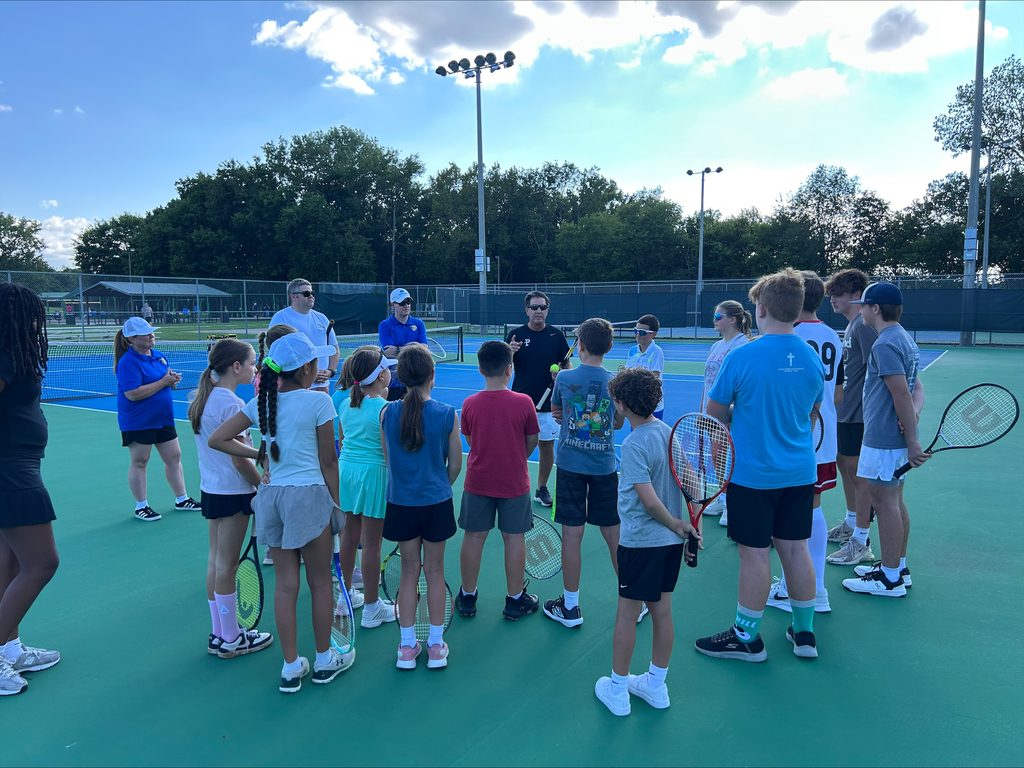 A group of children and adults on a tennis court, all holding tennis rackets. They are facing a tennis instructor.