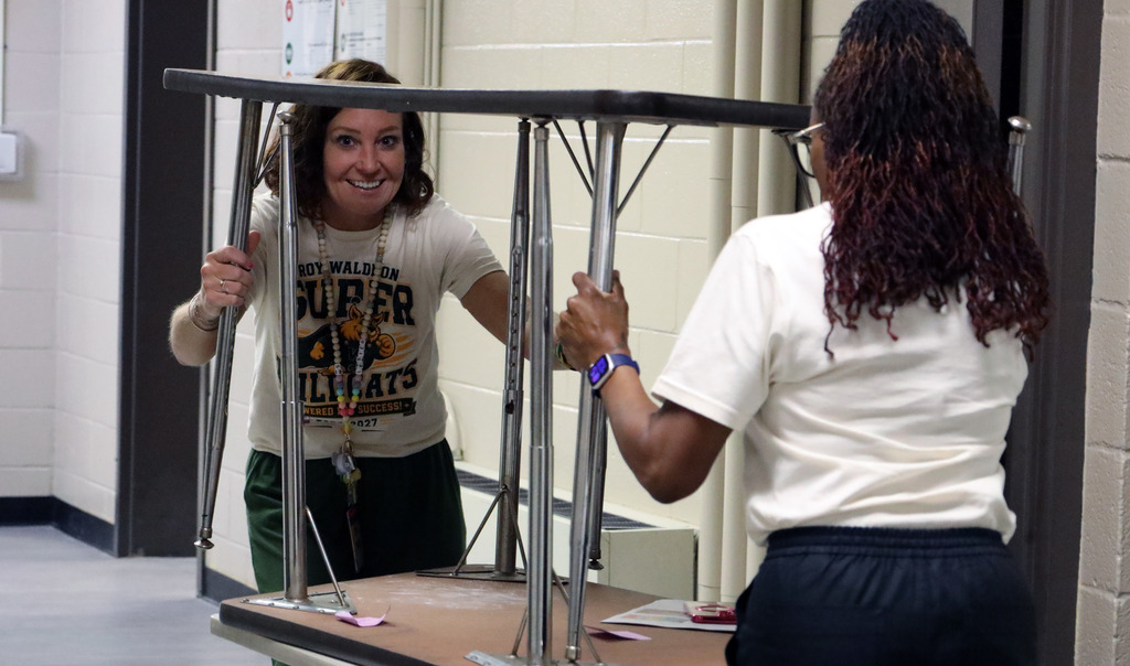 Two women in white tops and black pants hold up a folding table inside a room.