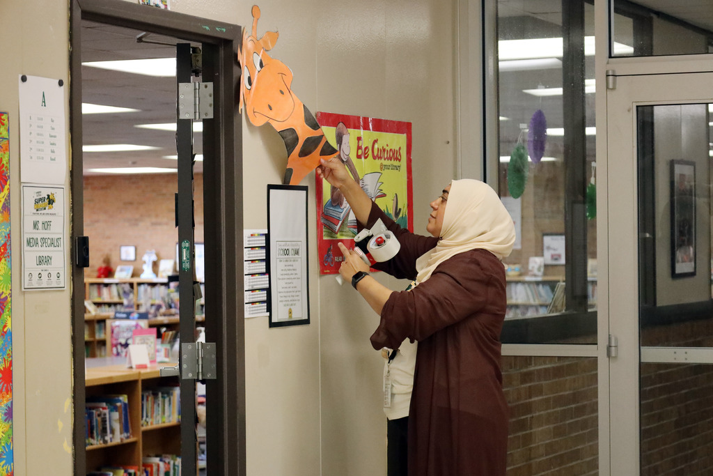A woman in a hijab and brown robe hangs a giraffe cutout on a bulletin board in a library.