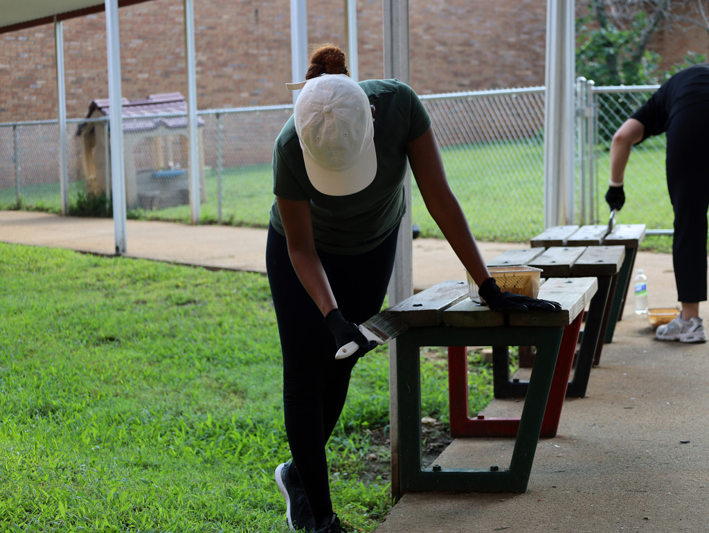 Two people clean benches on a sidewalk. One wears a white cap and gloves, the other a black shirt and pants.