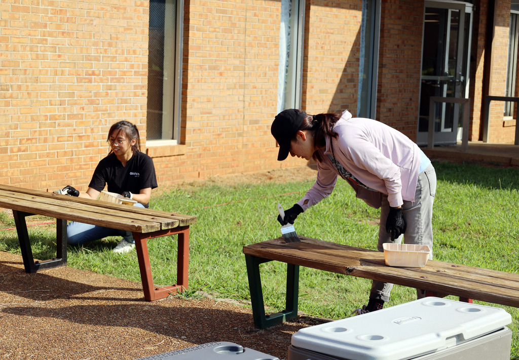 Two people in front of a brick building, one kneeling by a bench, the other standing by a bench painting.