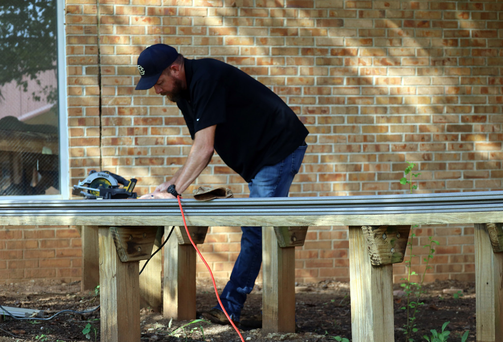A man wearing a black shirt and blue jeans uses a power tool on a wooden deck.
