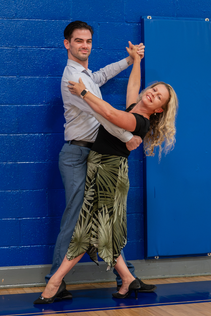 A man and woman dance close together in a gym with blue walls. The man is in a light blue shirt and the woman wears a black top.