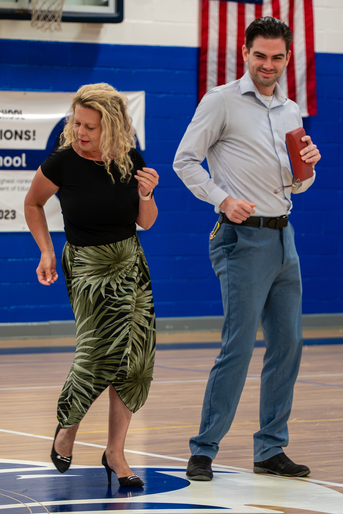 A woman and a man dance in an indoor court, wearing black and blue clothes. The background includes blue walls and an American flag.