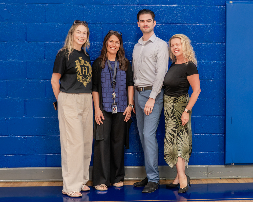 Four adults standing together against a blue wall. Three women and one man, each wearing different casual clothing.