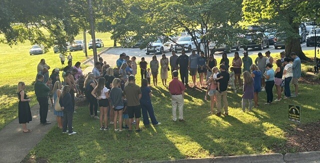 Group of people standing in an outdoor area, possibly in a park. Some wear sneakers. Cars parked.