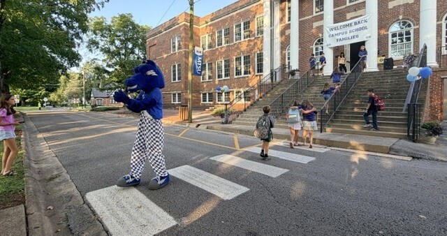 A school mascot stands at a crosswalk in front of a school building with steps and students walking.