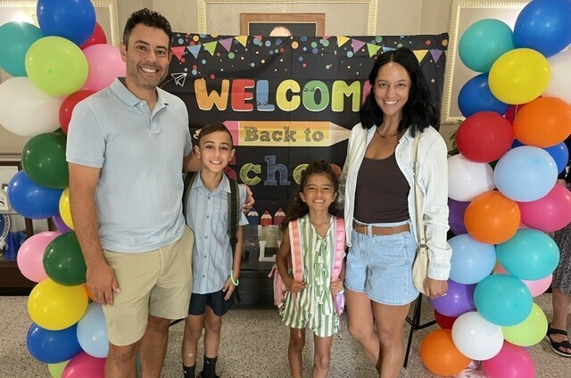 Family stands before a colorful balloon arch, with a "Welcome Back to School" sign.
