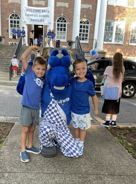 Three children, two in blue shirts, stand on a sidewalk with a mascot, in front of a building with a banner reading "Welcome Back Campus School Families."