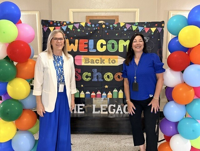 Two women stand before a colorful balloon arch and a chalkboard that reads "Welcome Back to School."