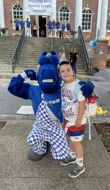 Child in school uniform stands with school mascot on a sidewalk in front of building.