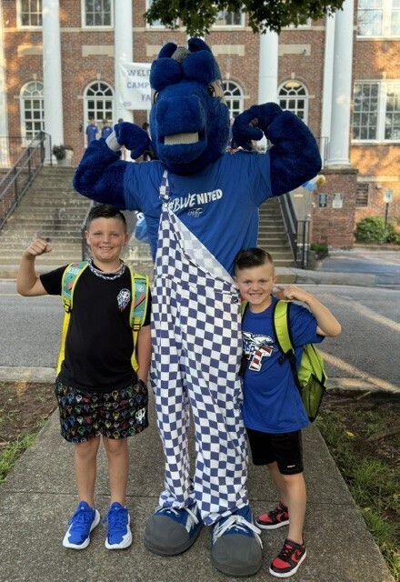 Two boys in school uniforms and backpacks stand next to a mascot with a blue outfit and backpack.