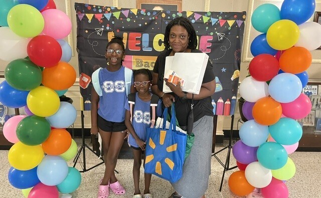 A woman stands with two children in front of a colorful balloon arch. They are near a blackboard with a "Welcome Back" banner.