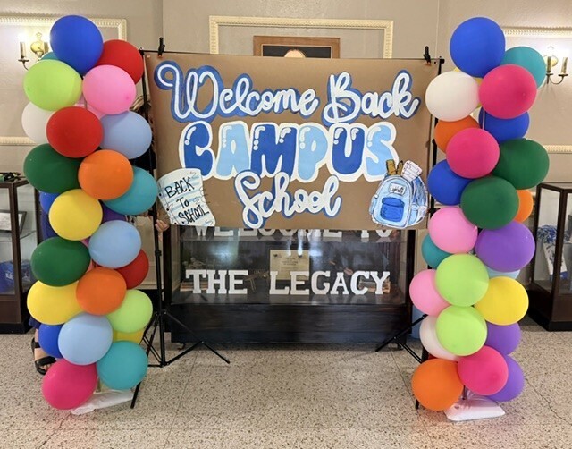 Colorful balloons create a cheerful entrance. A sign reads "Welcome Back Campus School The Legacy". Behind it, a black backdrop and glass displays.