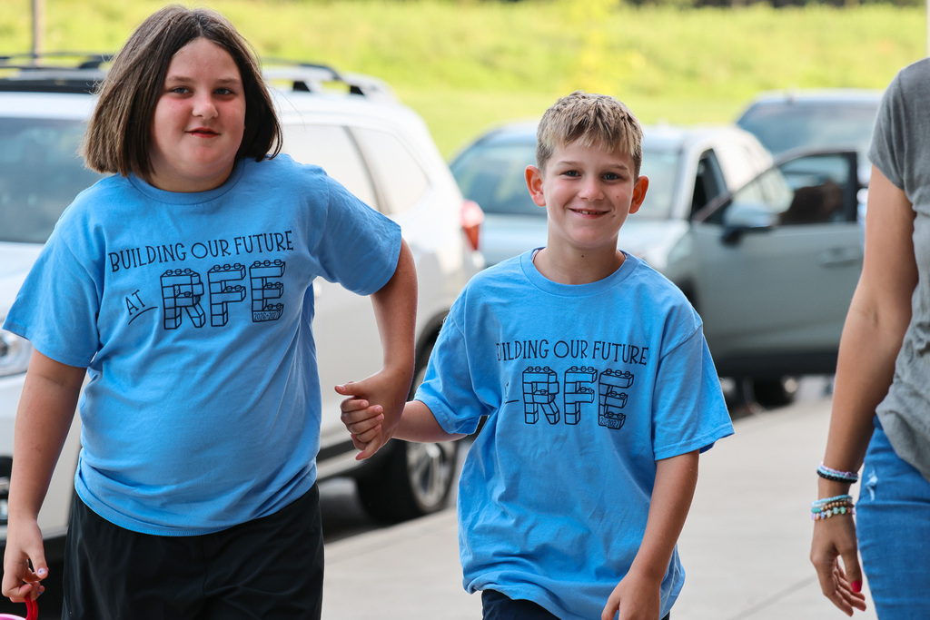 Two kids in matching blue T-shirts walk together on a sidewalk. Behind them are parked cars on a street.