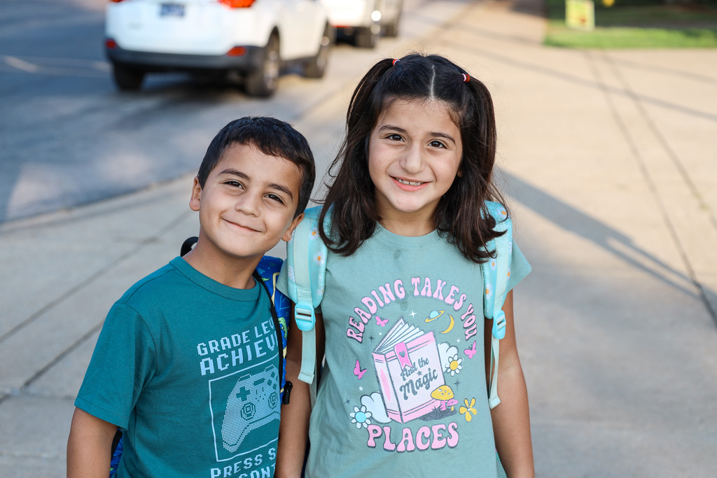 Two children stand on a sidewalk. The girl wears a light blue shirt with a book and text. The boy has a green shirt and a backpack.